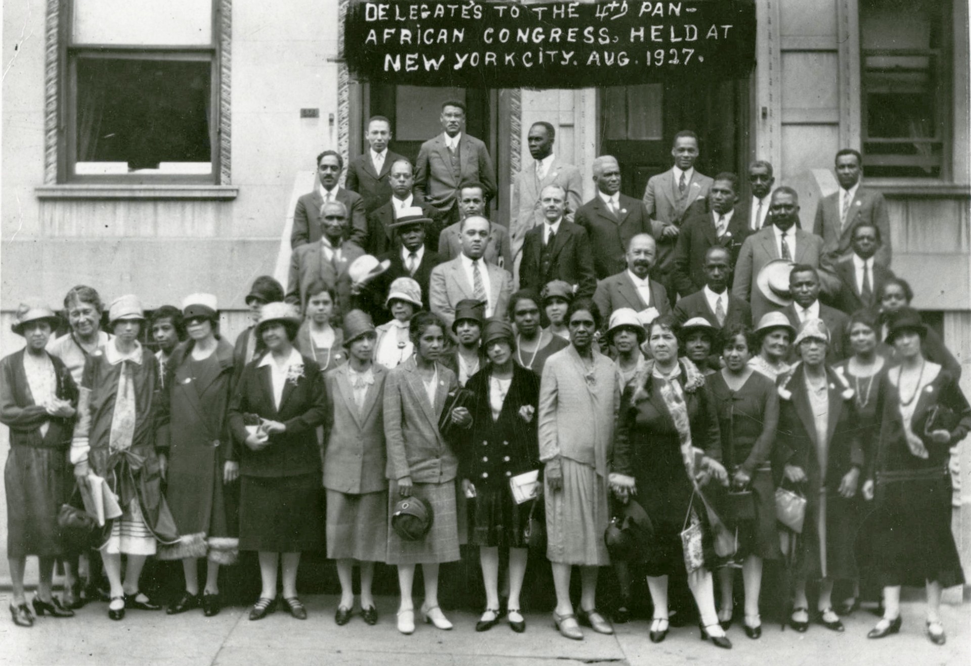Four rows of men and women in formal dress standing in a doorway with overlaid text at the top reading Delegates to the 4th Pan-African Congress. Held at New York City. Aug 1927.