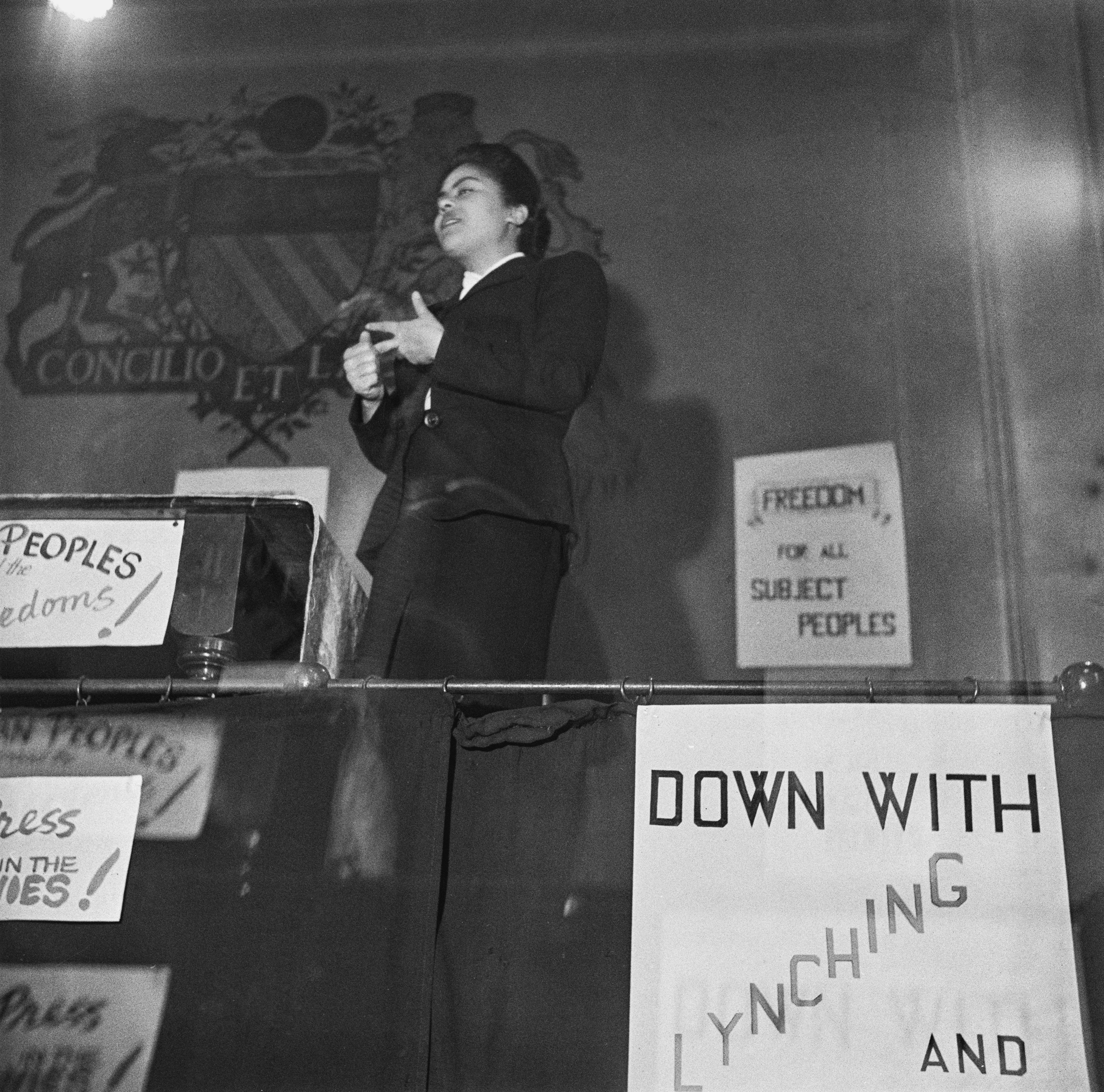 Black and white photo of woman speaking on stage surrounded by signs that say "Down with lynching" and "Freedom for all subject peoples"